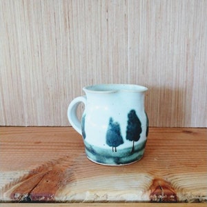 May include: A small white ceramic pitcher with a handle, featuring a hand-painted design of three dark green trees on a light green field.
