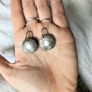 May include: A pair of silver earrings with a circular design. The earrings have a geometric pattern and are hanging from a silver hook.