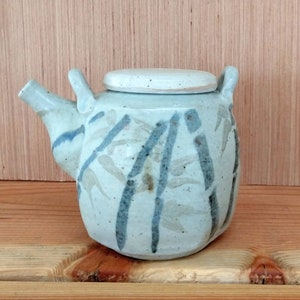 Puede incluir: Una tetera de cerámica blanca con tapa, con un diseño de bambú azul y gris. La tetera tiene un cuerpo redondeado y un pico en el lateral.