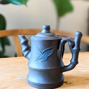 Puede incluir: Una tetera negra con un diseño inspirado en el bambú y un motivo de hoja azul. La tetera tiene un cuerpo redondeado y un pico que se curva hacia arriba. El asa también tiene forma de bambú.