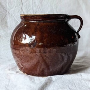 May include: A brown glazed ceramic crock with a single handle. The crock has a rounded body and a slightly flared rim.