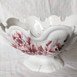 May include: A white ceramic bowl with a scalloped rim and a pedestal base. The bowl is decorated with a pink floral pattern.