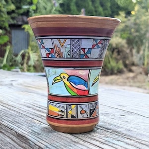 May include: A small, brown ceramic cup with a colorful design featuring a bird and geometric patterns. The cup is inscribed with the word "Aruba".
