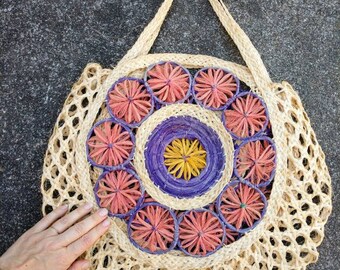 Può includere: Una borsa rotonda in paglia intrecciata con un design floreale viola, giallo e rosa. La borsa ha un'unica tracolla ed è realizzata con materiali naturali.
