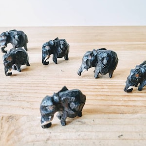 May include: Six small black wooden elephant figurines with ivory tusks. The elephants are arranged in a semi-circle on a wooden surface.