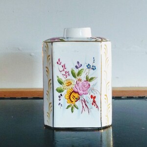May include: A white porcelain jar with a gold trim and a floral design. The flowers are pink, yellow, and blue.