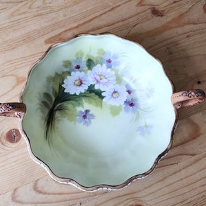 May include: A white porcelain bowl with two handles. The bowl has a scalloped edge and is decorated with a floral design of white and purple flowers on a green background.