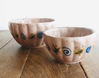 May include: Two antique, light pink ceramic bowls with a fluted design. Each bowl features hand-painted blue, green, and yellow spiral and leaf motifs. The bowls are displayed on a wooden surface.