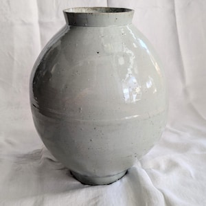 Może przedstawiać: Jasnoszary ceramiczny wazon o zaokrąglonym korpusie i wąskiej szyi. Wazon ma gładkie, błyszczące wykończenie i stoi na białej tkaninie. Wazon jest przedmiotem dekoracyjnym.