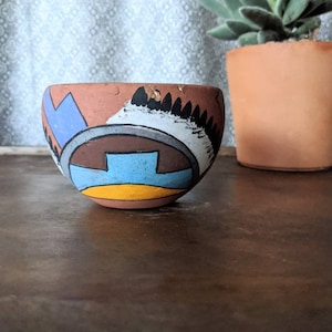 May include: A small, hand-painted terracotta bowl with a geometric design in shades of blue, yellow, brown, and white. The bowl is sitting on a wooden surface.
