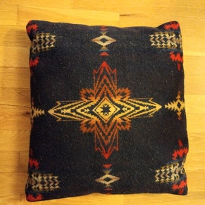 Op de afbeelding: Een zwart kussen met een kleurrijk geometrisch patroon in tinten rood, oranje, geel en wit. Het patroon doet denken aan Native American kunst.