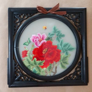 Pode incluir: Uma moldura de madeira preta com um desenho circular bordado de peónias rosa e vermelhas com folhas verdes sobre um fundo branco.