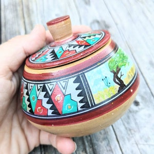 May include: A small, round, ceramic container with a lid. The container is decorated with a colorful geometric pattern and a painted scene of a tree and flowers. The lid is also decorated with a geometric pattern.