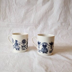 May include: Two white ceramic mugs with blue illustrations of figures and floral designs. The mugs have a cylindrical shape with a handle on the side. The illustrations are in a folk art style.