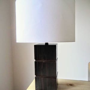 May include: A dark brown wooden table lamp with a white fabric lampshade. The lamp base is made of three stacked square blocks.