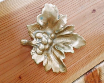 May include: A gold-colored metal grapevine leaf design with a raised grape cluster in the center. The leaf is detailed with intricate carvings and has a slightly aged patina.