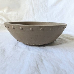 May include: A large, oval-shaped, light brown bowl with a series of small, evenly spaced, raised dots around the rim and base. The bowl has a smooth, matte finish and a simple, elegant design. The bowl is likely made of ceramic or a similar material.