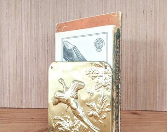May include: A brass bookend with an embossed design of a pheasant in flight. The bookend is holding up two books, one of which has the text "Woodcraft" on the spine.