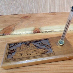 Op de afbeelding: Een houten pennenhouder met een gegraveerd beeld van een schip en wolken. De houder heeft een doorzichtige plastic pen erin gestoken.