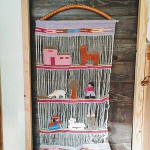 May include: A woven wall hanging with a light purple background and a brown wooden hanger. The hanging features a variety of colorful animals and houses, including a llama, a sheep, and a small house with a pink roof.
