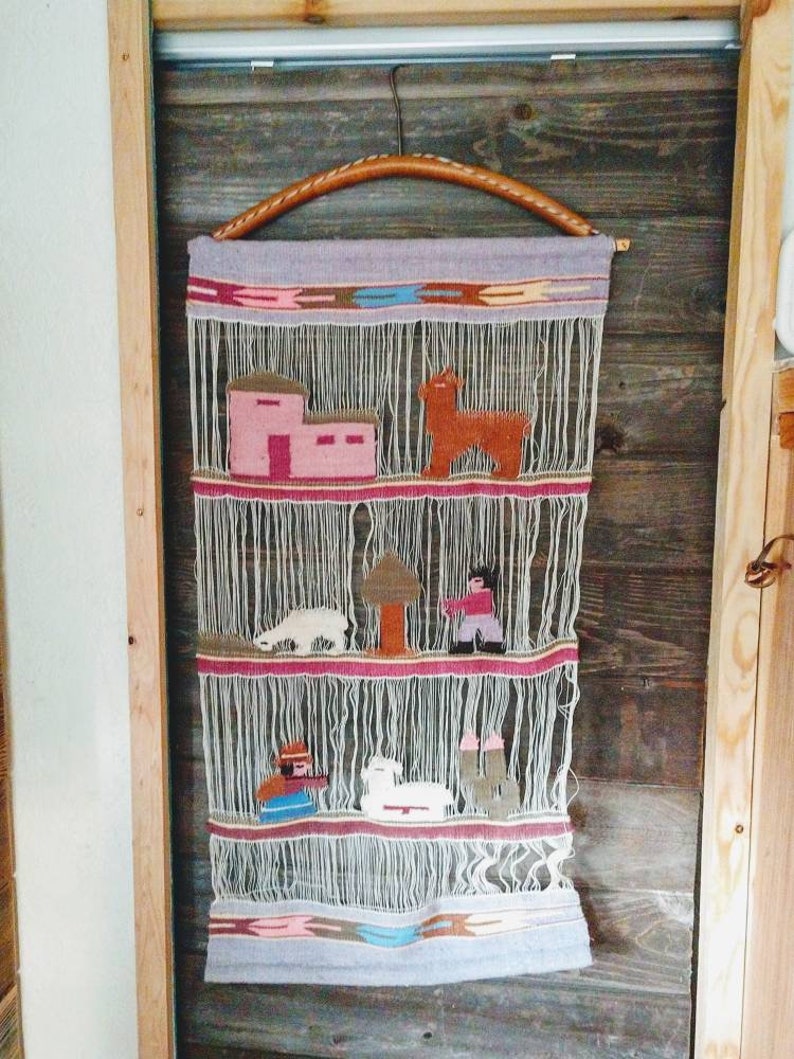 May include: A woven wall hanging with a light purple background and a brown wooden hanger. The hanging features a variety of colorful animals and houses, including a llama, a sheep, and a small house with a pink roof.