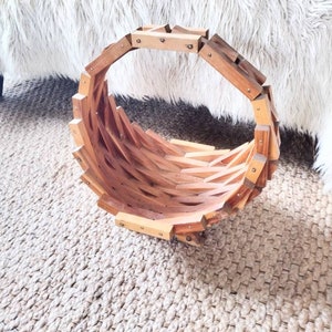 May include: A handcrafted wooden basket with a unique, geometric design. The basket is constructed from light-colored wood and features a circular shape with a woven interior. The basket is approximately 12 inches in diameter and is suitable for decorative or storage purposes.