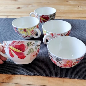 May include: Set of five floral teacups with a white background and colorful floral designs. The cups are arranged in a semi-circle on a black and white patterned surface.