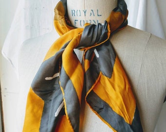 May include: A yellow and grey patterned silk scarf tied around a mannequin's neck. The scarf has a geometric design with white accents.