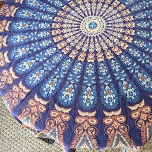 Puede incluir: Un tapiz redondo con un diseño de mandala en tonos azules, naranjas y blancos. El tapiz tiene un fondo morado oscuro y presenta patrones intrincados.