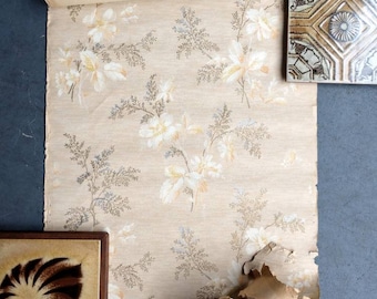 May include: A roll of vintage wallpaper with a floral pattern featuring white flowers and brown branches on a beige background. The wallpaper is partially unrolled and has a slightly distressed look.