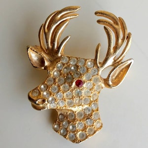 May include: A gold-toned reindeer brooch with a red rhinestone for the nose and clear rhinestones covering the rest of the reindeer's head and antlers.