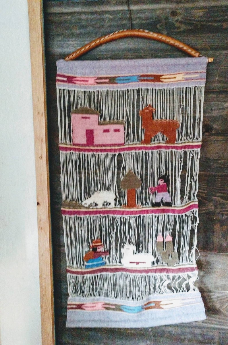 May include: A woven wall hanging with a light gray background and colorful geometric patterns. The hanging features various figures, including a house, a tree, and animals, all woven into the fabric.