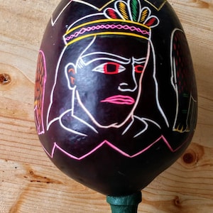 May include: A black gourd maraca with a painted design of a person wearing a crown and feathers. The person has red eyes and pink lips. The maraca has a green wooden handle.