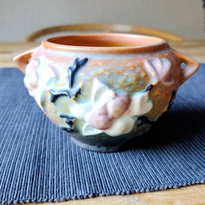 Puede incluir: Un pequeño cuenco de cerámica marrón con un diseño floral. El cuenco tiene dos asas y está decorado con flores blancas y rosas y tallos negros.