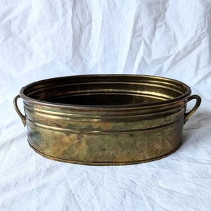 May include: An oval brass planter with two handles. The planter has a worn, aged look and is made of a thick metal.