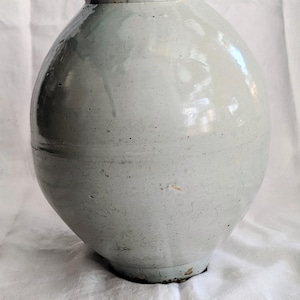 May include: A large, pale blue ceramic vase with a rounded body and a slightly flared neck. The vase has a smooth, glossy finish and sits on a white cloth. The vase is approximately 12 inches tall.
