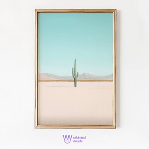 Può includere: Una stampa incorniciata di un singolo cactus in un paesaggio desertico. Il cielo è azzurro chiaro e la sabbia è rosa pallido. Il cactus è verde e ha alcuni rami.