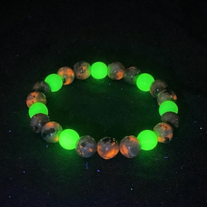 May include: A circular beaded bracelet featuring alternating spherical beads. Some beads are dark gray with orange mottled patterns, while others are solid, bright neon green and emit a distinct glow against the dark environment.