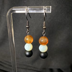 Puede incluir: Un par de pendientes colgantes con un diseño de tres cuentas. Cada pendiente presenta una cuenta naranja en la parte superior, una cuenta blanca en el medio y una cuenta negra en la parte inferior. Las cuentas están suspendidas de un gancho plateado.