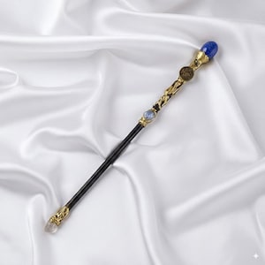 May include: An ornate wand with a black shaft and gold detailing. It features a blue gemstone at the top, a clear crystal at the base, and further gemstones and a circular emblem along the shaft. The wand is displayed on a white satin cloth.