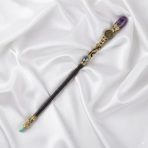 May include: A decorative wand with a dark wooden shaft and ornate gold-coloured metal accents. The wand features a purple gemstone at the top, a blue gemstone in the middle, and a green gemstone at the base. The wand is approximately 35.5 cm long.