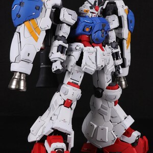 Puede incluir: Un modelo articulado y detallado de un robot Gundam, principalmente blanco con detalles en azul, rojo y amarillo. El modelo presenta grandes protectores de hombros, propulsores y l&iacute;neas de paneles intrincadas.