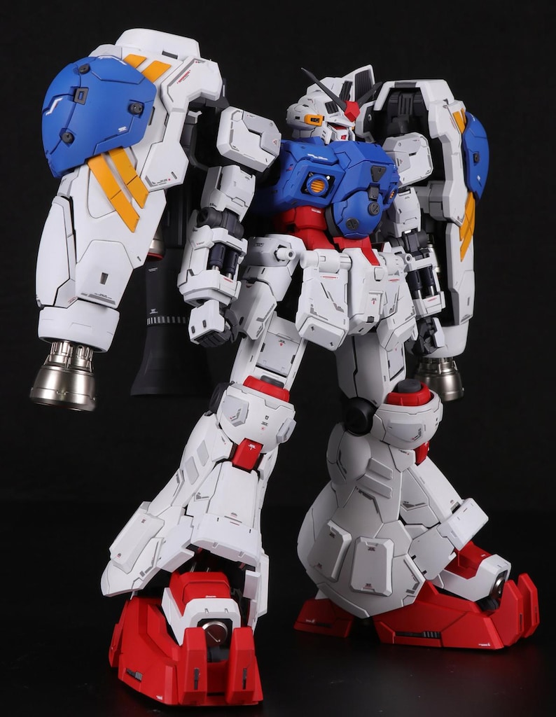 Puede incluir: Un modelo articulado y detallado de un robot Gundam, principalmente blanco con detalles en azul, rojo y amarillo. El modelo presenta grandes protectores de hombros, propulsores y l&iacute;neas de paneles intrincadas.