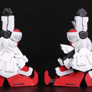 Puede incluir: Dos componentes de piernas rob&oacute;ticas blancas y rojas, posiblemente de un kit de modelo, se colocan uno al lado del otro sobre un fondo negro. Los componentes presentan paneles y detalles intrincados, con detalles en rojo en los pies y las articulaciones.