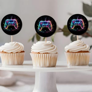 May include: Three cupcakes decorated with colourful, neon-style video game controller toppers. The cupcakes are white with white frosting and are displayed on a white cake stand.