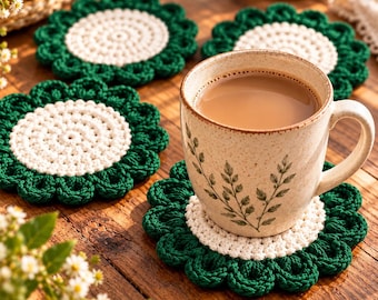 May include: A ceramic mug with a green leaf pattern sits on a round, flower-shaped crochet coaster. The coaster features a white circular center surrounded by dark green scalloped petals. Three additional identical coasters are arranged nearby.