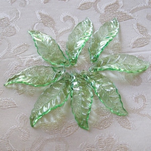 May include: A collection of eight green glass leaves with a textured surface. The leaves are arranged in a circular pattern on a white fabric background.