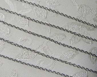 May include: Four strands of black chain on a white fabric background. The chain is made of small, linked metal rings.