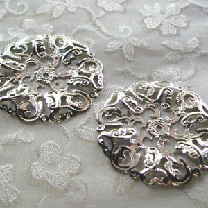 May include: Two silver filigree metal embellishments with a circular design. The embellishments have a delicate, intricate pattern of swirls and curves.