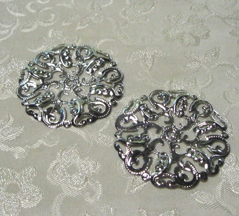 May include: Two silver filigree metal embellishments with a circular design. The embellishments have a delicate, intricate pattern.